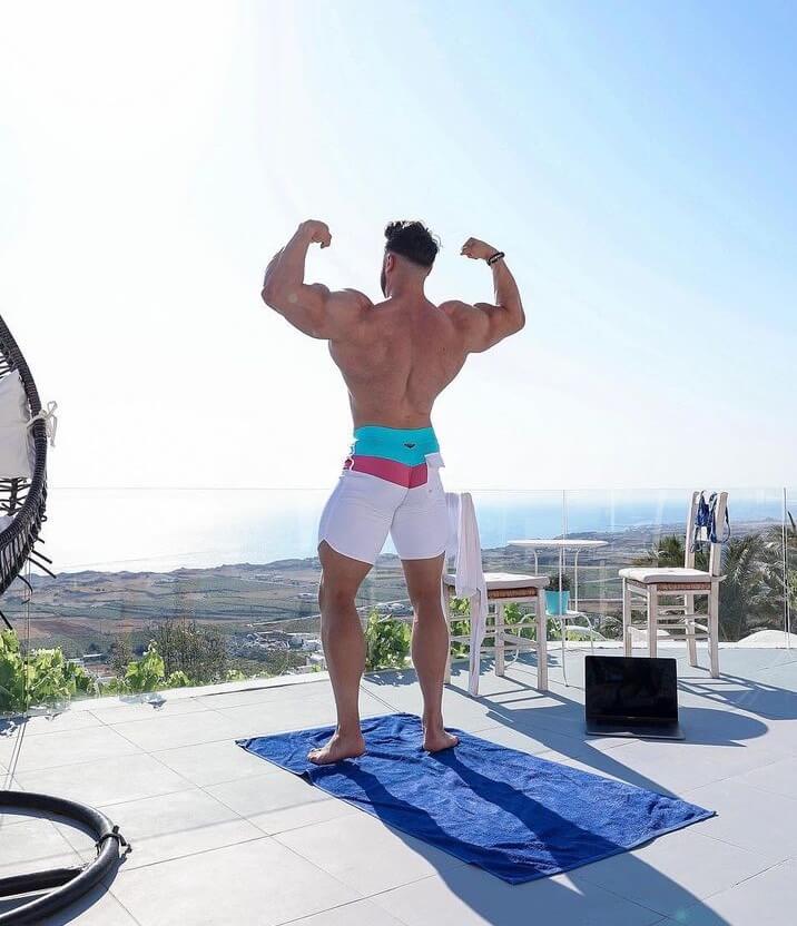 Brandon Harding doing a front double biceps pose while shirtless outside with a great view.